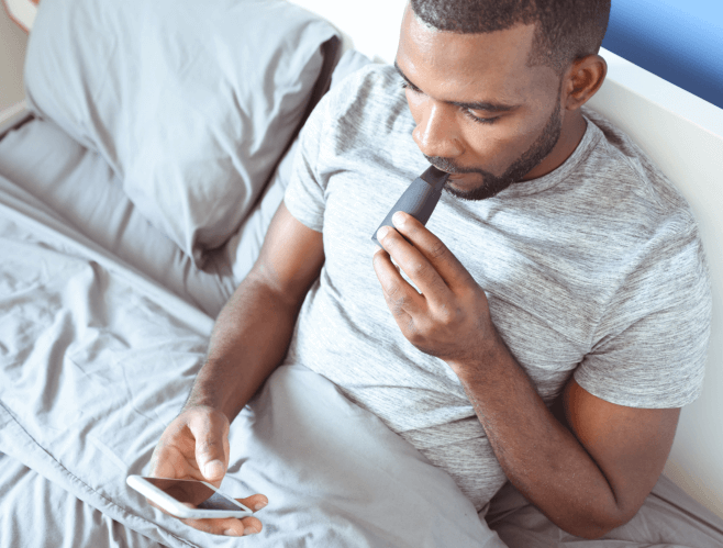 A man in bed blows into a Lumen device and holds a cell phone in his hand